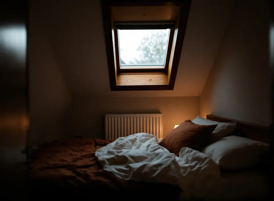Een zolderslaapkamer onder een schuin dak met een dakraam, een lage radiator en een bed met roestbruin beddengoed