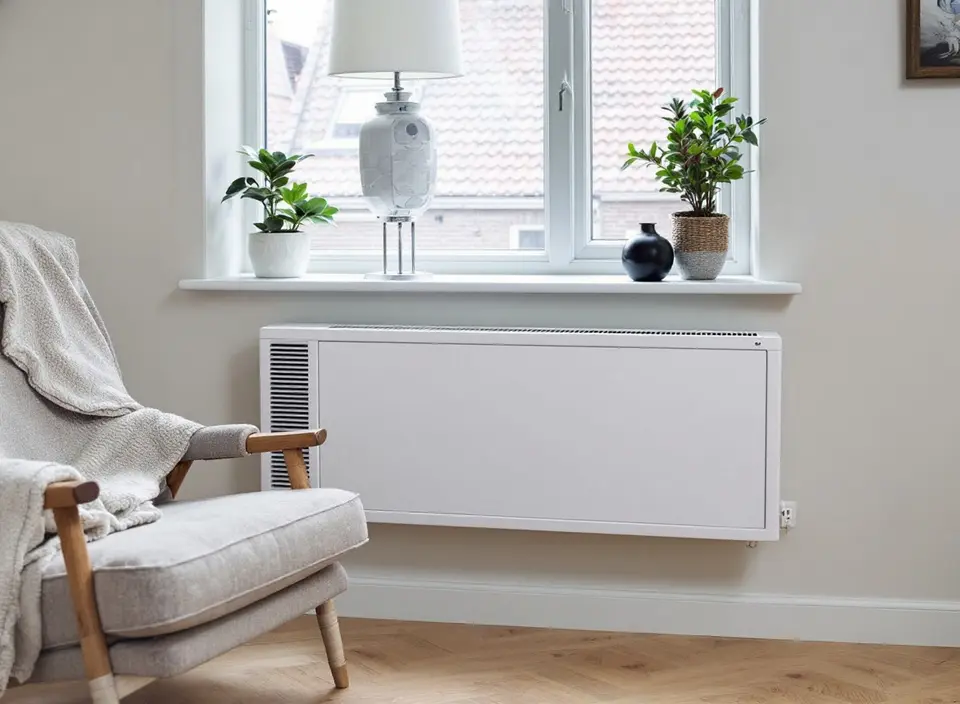 Een lage witte convector met ventilator en een rooster aan de zijkant onder een raam, met een fauteuil ernaast